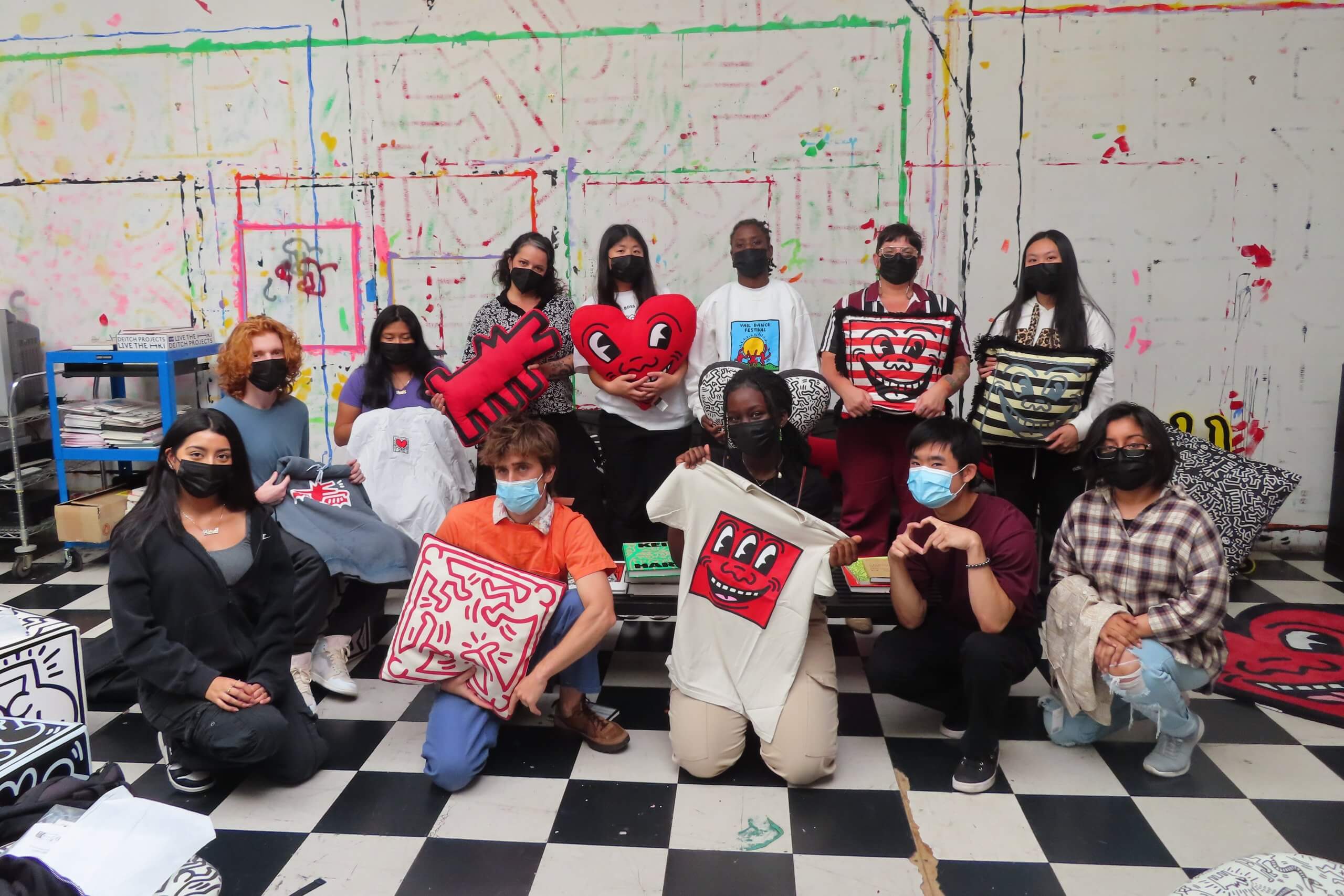 A group of 12, half standing behind a black couch and half crouching in front on a black and white checkered floor pose for the camera in a space that was once the studio of the artist Keith Haring.